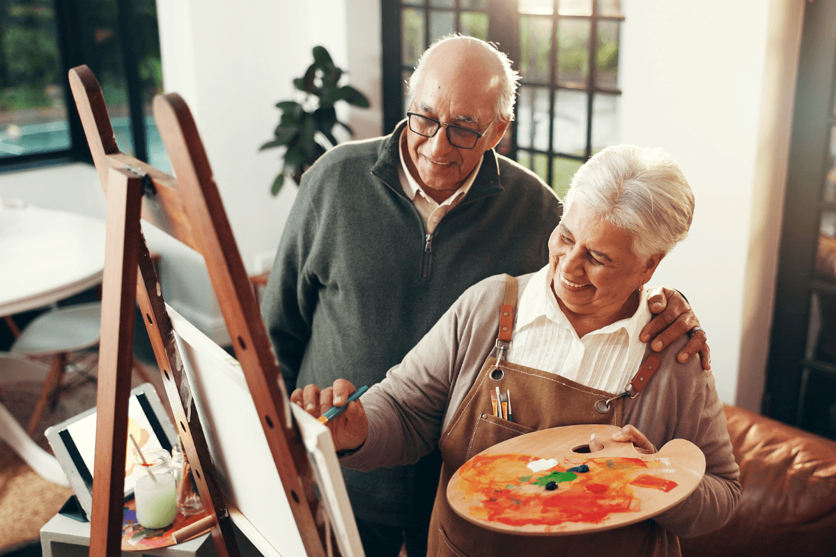 A smiling senior couple shares a joyful moment as they paint and sketch together on canvas.